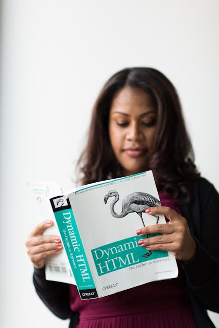 A woman with dark brown hair, dressed in a black blazer and maroon top, is holding an open technical book titled 'Dynamic HTML' with both hands, examining its content. The book has a predominantly whi