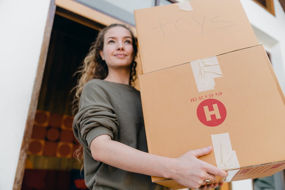 A young woman with curly hair, dressed in a grey long-sleeved top, is holding two large cardboard boxes during a home relocation. One box is marked with the word 'toys' written in pencil on the top. S
